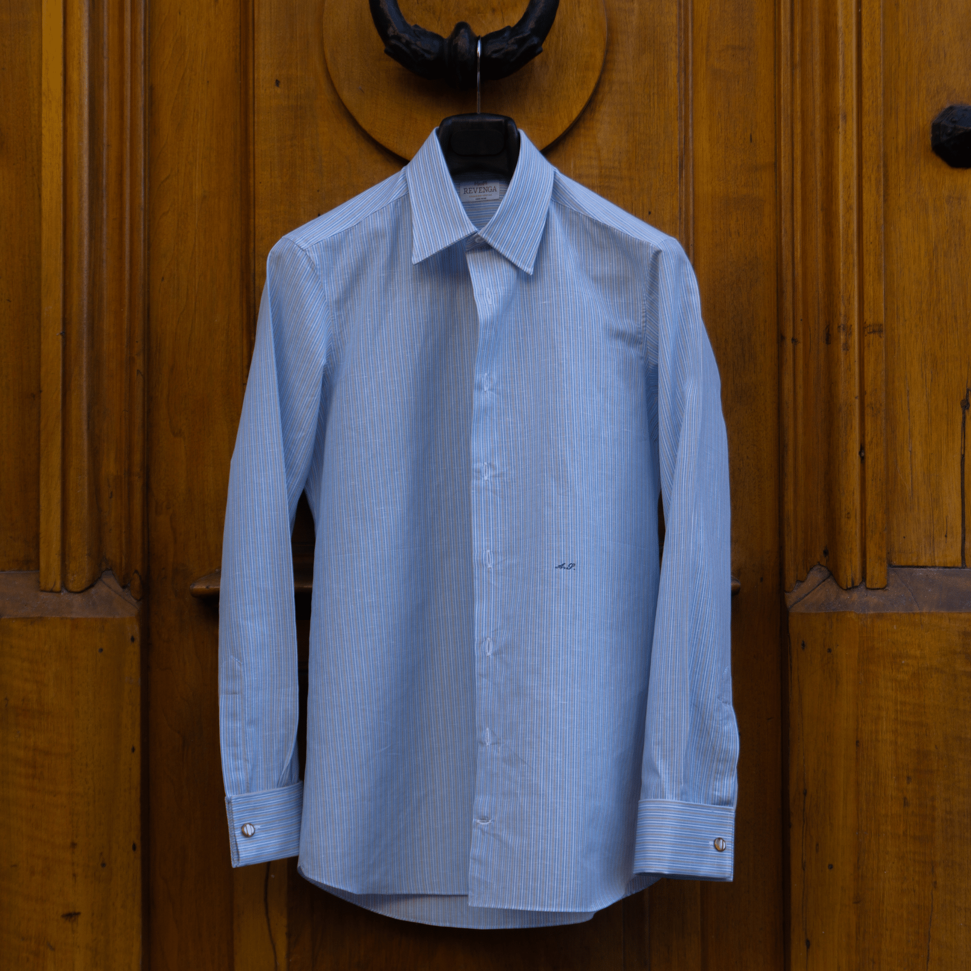 Vue d'ensemble d'une chemise sur mesure réalisée par le tailleur genevois Revenga. 

La chemise est sur un portant. Ce dernier est accroché à une belle porte en bois blond. 

Il s'agit d'une chemise sur mesure réalisée en tissu Zephirlino de la maison Alumo.

Ce tissu est composé de lin et de coton, il est rayé bleu, blanc et beige.